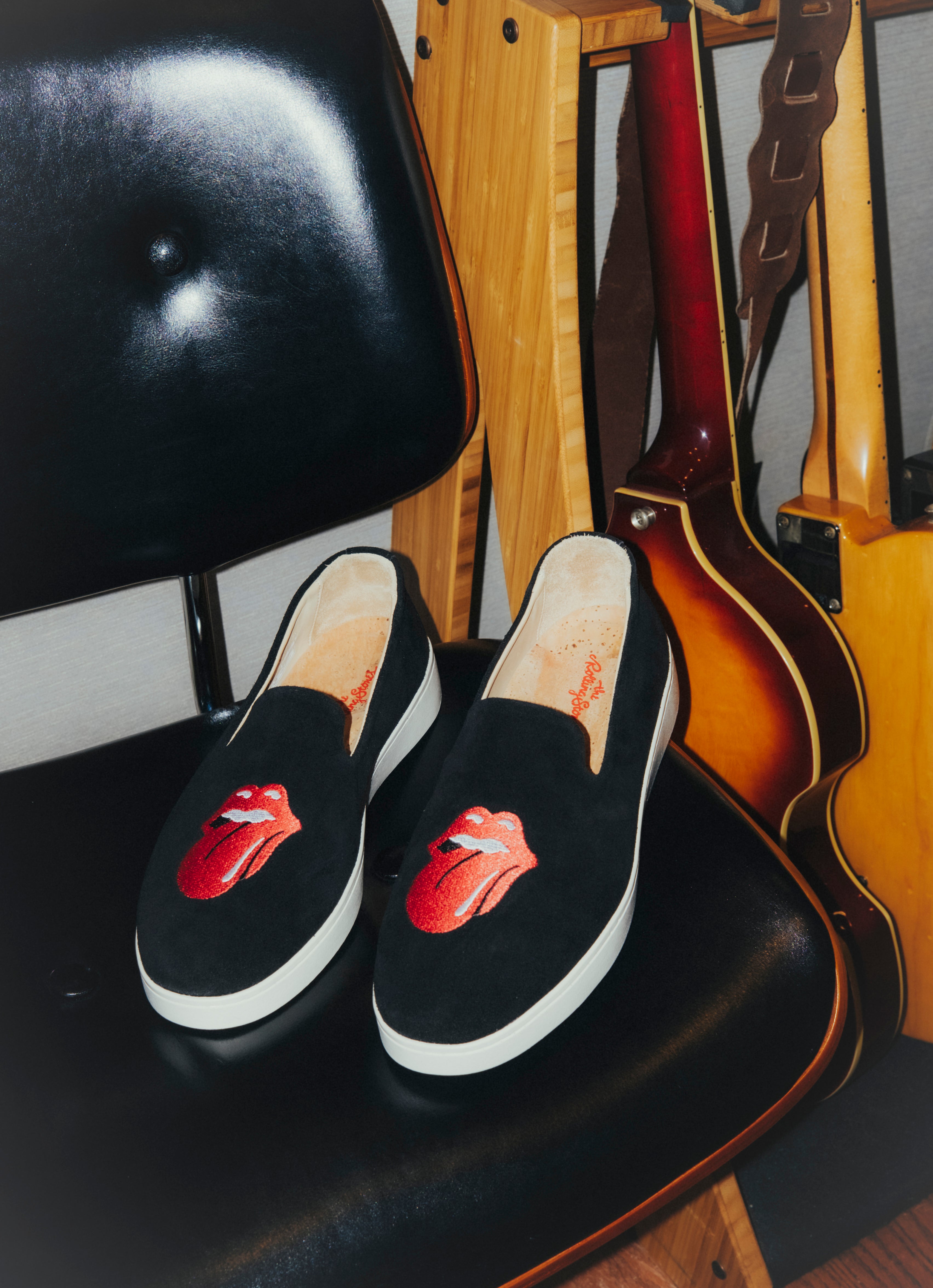 Black slip-on shoes with red lips design on a chair with guitars in the background The Rolling Stones 